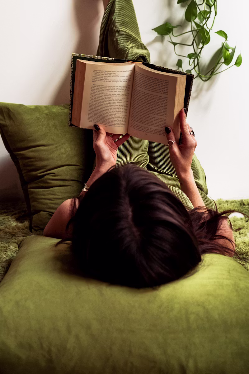 A woman resting on olive-green cushions reading a book, at ease in a calm space